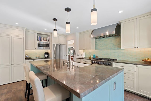 kitchen with an island with sink, appliances with stainless steel finishes, pendant lighting, and wall chimney exhaust hood