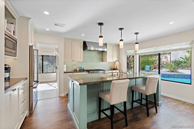 kitchen featuring appliances with stainless steel finishes, a kitchen island with sink, wall chimney range hood, and a kitchen breakfast bar