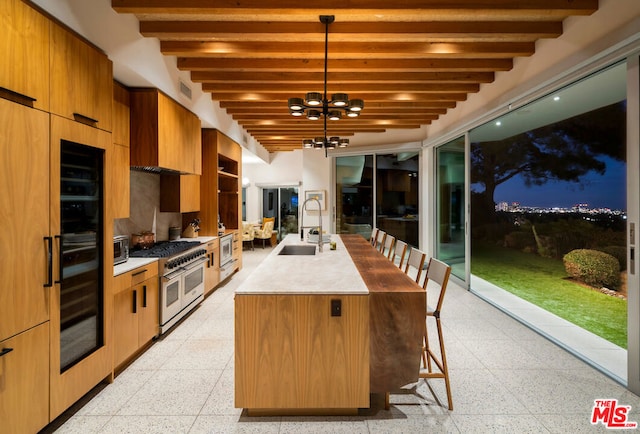 kitchen with hanging light fixtures, a notable chandelier, an island with sink, beamed ceiling, and range with two ovens