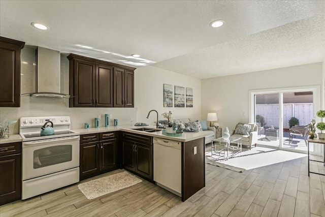 kitchen with sink, wall chimney range hood, white appliances, dark brown cabinetry, and kitchen peninsula