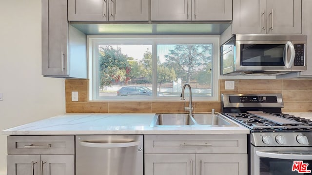 kitchen with sink, light stone counters, gray cabinets, stainless steel appliances, and decorative backsplash