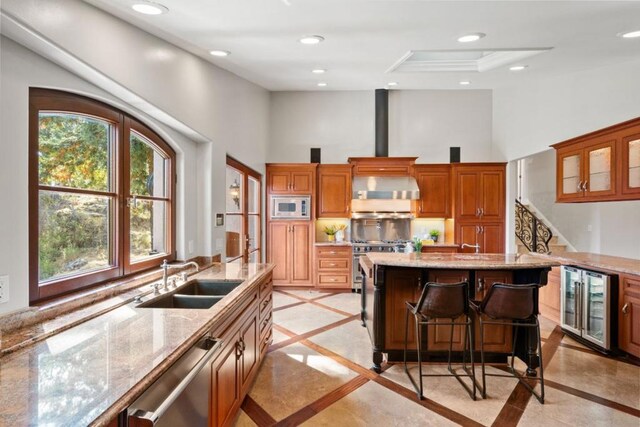 kitchen with wall chimney exhaust hood, sink, a center island with sink, appliances with stainless steel finishes, and beverage cooler