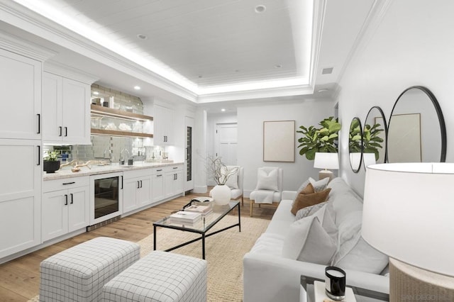 living room featuring beverage cooler, ornamental molding, a tray ceiling, wet bar, and light hardwood / wood-style flooring