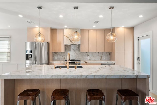 kitchen with a kitchen bar, light brown cabinetry, hanging light fixtures, stainless steel fridge, and light stone countertops