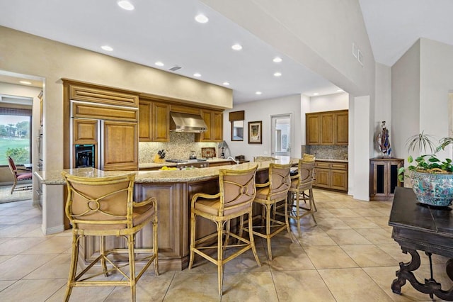 kitchen featuring a large island with sink, light tile patterned floors, a kitchen breakfast bar, light stone countertops, and wall chimney range hood