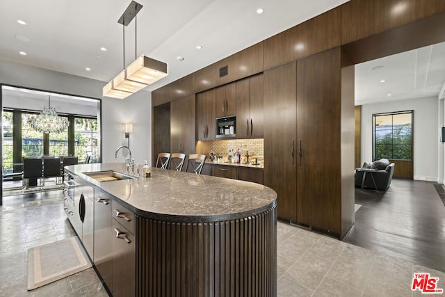 kitchen featuring sink, backsplash, hanging light fixtures, a notable chandelier, and a center island with sink