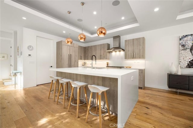 kitchen with sink, decorative light fixtures, light wood-type flooring, a raised ceiling, and wall chimney range hood