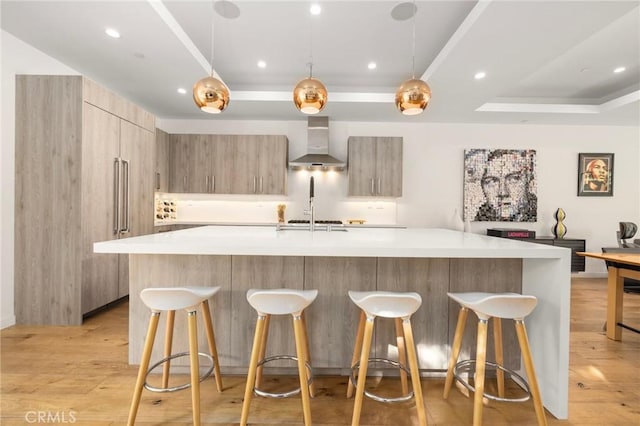 kitchen featuring decorative light fixtures, a tray ceiling, a kitchen island with sink, light hardwood / wood-style floors, and wall chimney range hood