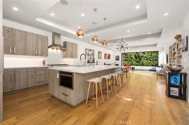 kitchen with hanging light fixtures, wall chimney range hood, a large island with sink, and a tray ceiling