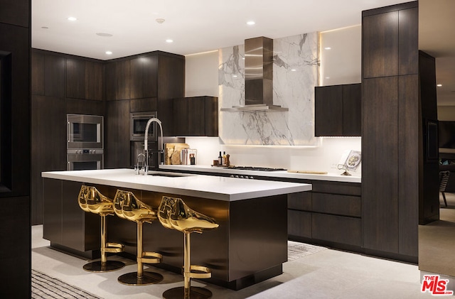 kitchen featuring appliances with stainless steel finishes, a breakfast bar, sink, a center island with sink, and wall chimney exhaust hood