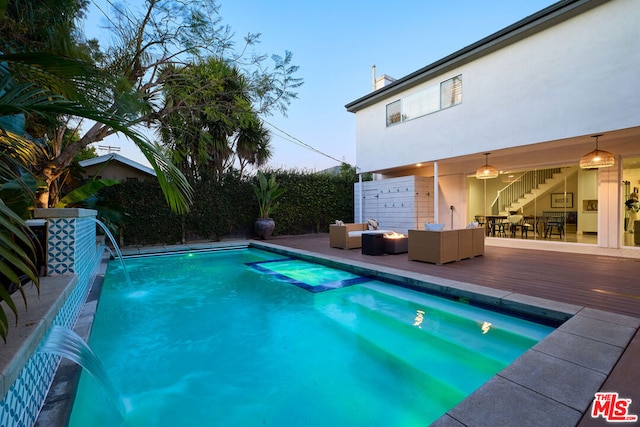 pool at dusk featuring a wooden deck, an outdoor living space with a fire pit, and pool water feature