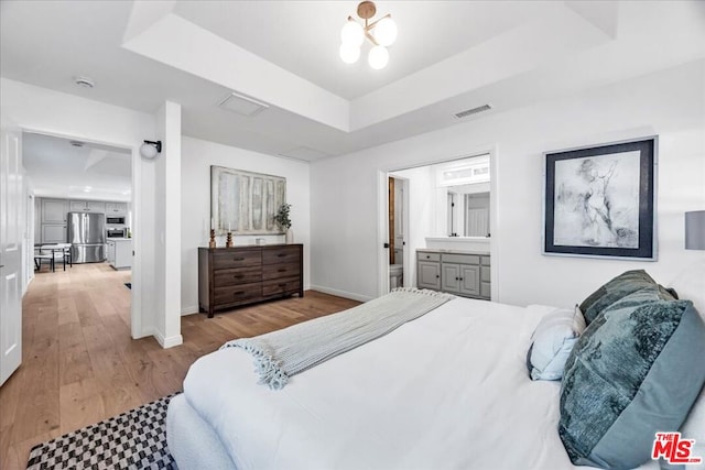 bedroom with connected bathroom, stainless steel fridge, a raised ceiling, and light wood-type flooring