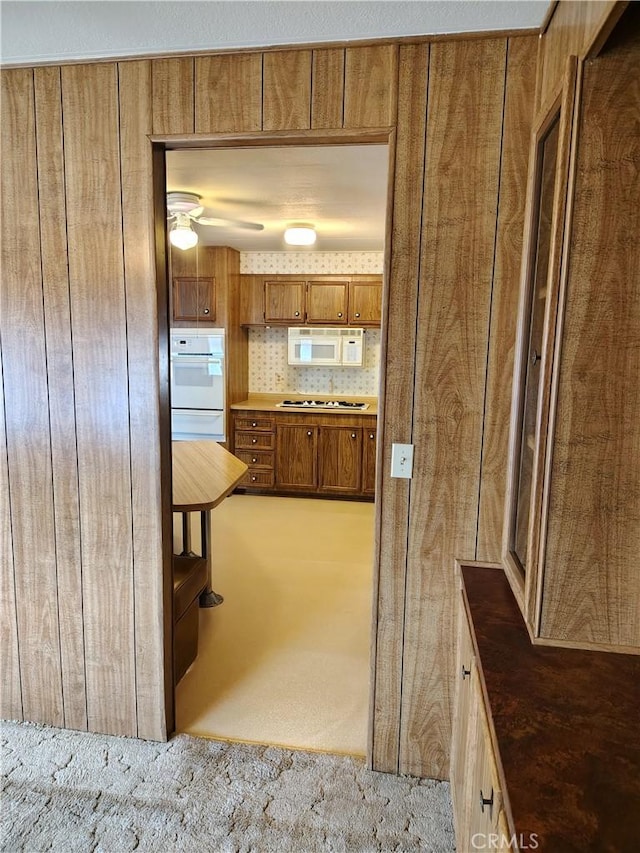 kitchen featuring brown cabinets, a warming drawer, dark countertops, light colored carpet, and white appliances