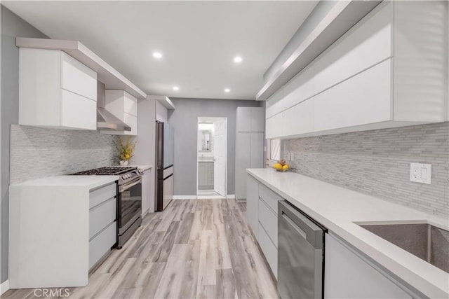 kitchen with tasteful backsplash, white cabinetry, stainless steel appliances, wall chimney range hood, and light hardwood / wood-style flooring