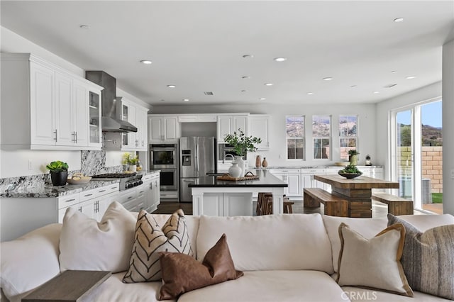 kitchen with a kitchen island, white cabinets, dark stone counters, stainless steel appliances, and wall chimney range hood