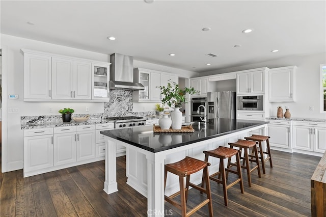 kitchen featuring wall chimney exhaust hood, a kitchen bar, a center island with sink, appliances with stainless steel finishes, and white cabinets