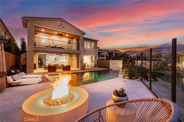 back house at dusk featuring a fenced in pool, ceiling fan, a patio, an outdoor living space with a fire pit, and a balcony