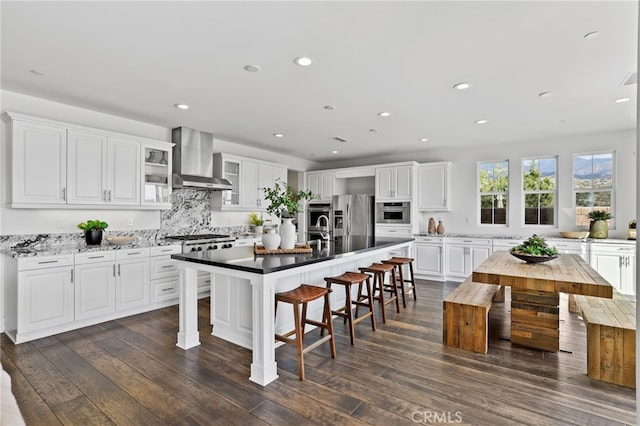 kitchen with wall chimney range hood, dark wood-type flooring, white cabinetry, a kitchen island with sink, and stainless steel refrigerator with ice dispenser