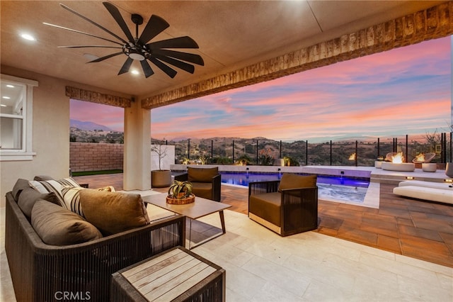 patio terrace at dusk with ceiling fan, an outdoor living space with a fire pit, a fenced in pool, and a mountain view