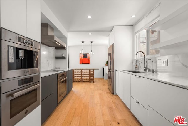 kitchen with sink, decorative light fixtures, white cabinets, stainless steel appliances, and wall chimney range hood