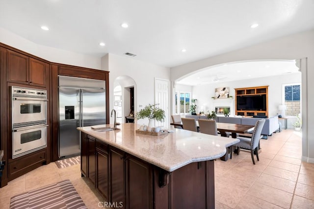 kitchen featuring sink, white double oven, light stone countertops, an island with sink, and built in fridge