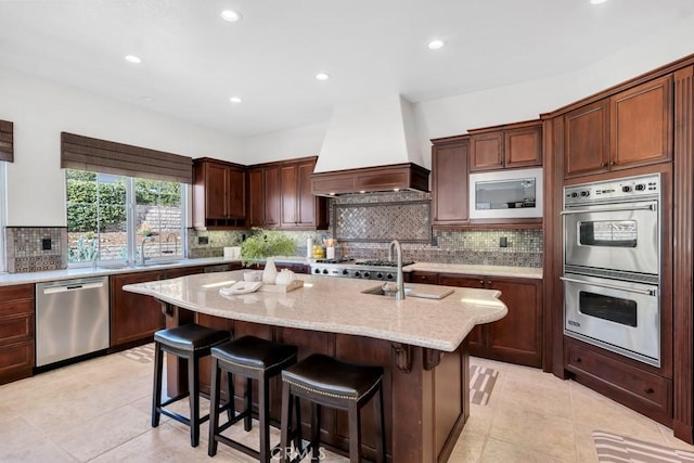 kitchen with appliances with stainless steel finishes, a kitchen island with sink, light stone countertops, a kitchen bar, and custom exhaust hood