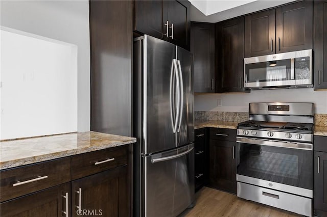 kitchen featuring stainless steel appliances, dark brown cabinets, light stone counters, and light wood-type flooring