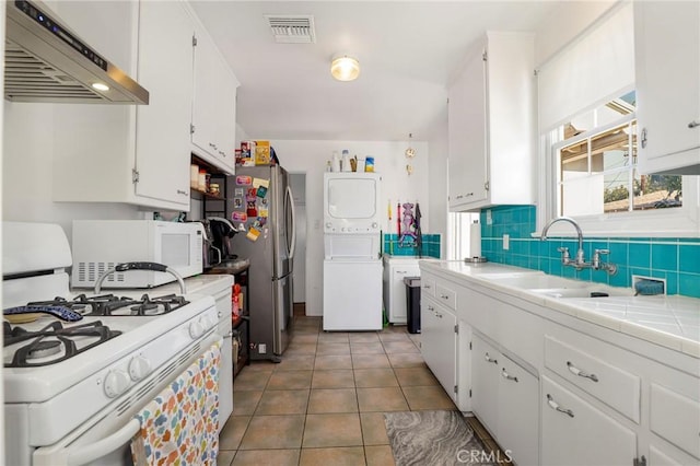 kitchen featuring stacked washer / dryer, white cabinetry, exhaust hood, tile counters, and white appliances