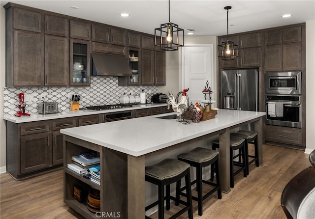 kitchen with dark brown cabinetry, stainless steel appliances, a sink, ventilation hood, and open shelves