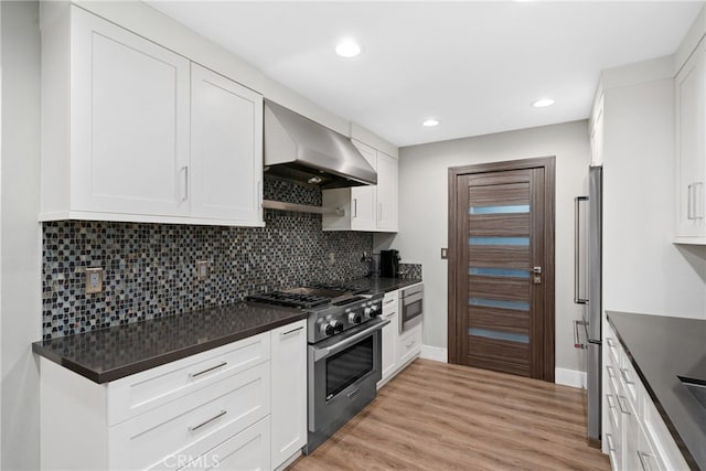 kitchen with tasteful backsplash, appliances with stainless steel finishes, wall chimney range hood, light hardwood / wood-style floors, and white cabinets