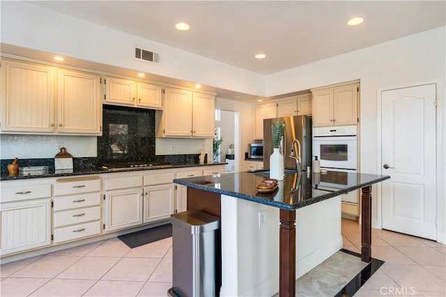 kitchen with a kitchen island with sink, light tile patterned floors, dark stone counters, and appliances with stainless steel finishes