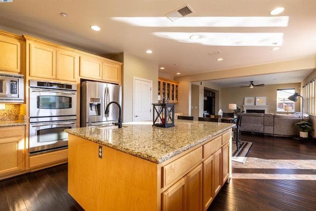 kitchen with light stone counters, appliances with stainless steel finishes, dark hardwood / wood-style floors, an island with sink, and ceiling fan