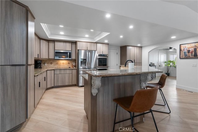 kitchen featuring a kitchen breakfast bar, stainless steel appliances, light stone countertops, a center island with sink, and a raised ceiling