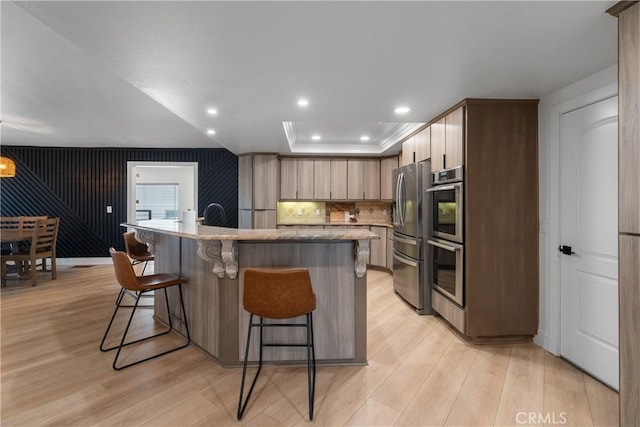 kitchen with light wood-type flooring, appliances with stainless steel finishes, a kitchen breakfast bar, a raised ceiling, and a kitchen island with sink