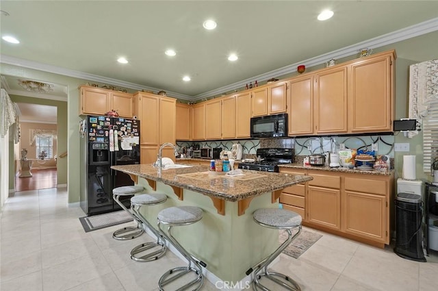 kitchen with light stone counters, light brown cabinetry, a center island with sink, and black appliances