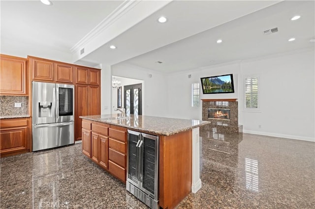 kitchen with crown molding, stainless steel fridge, a kitchen island with sink, light stone countertops, and beverage cooler