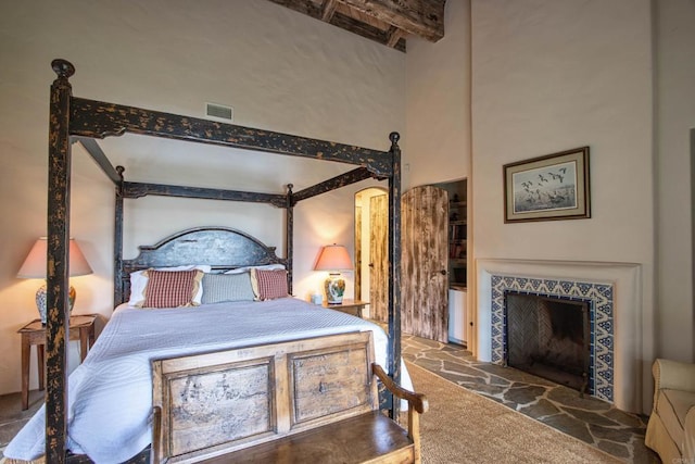 bedroom with a fireplace with raised hearth, visible vents, a high ceiling, stone finish floor, and beamed ceiling