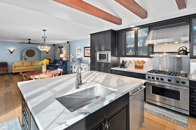 kitchen with backsplash, appliances with stainless steel finishes, a sink, wall chimney range hood, and dark cabinetry