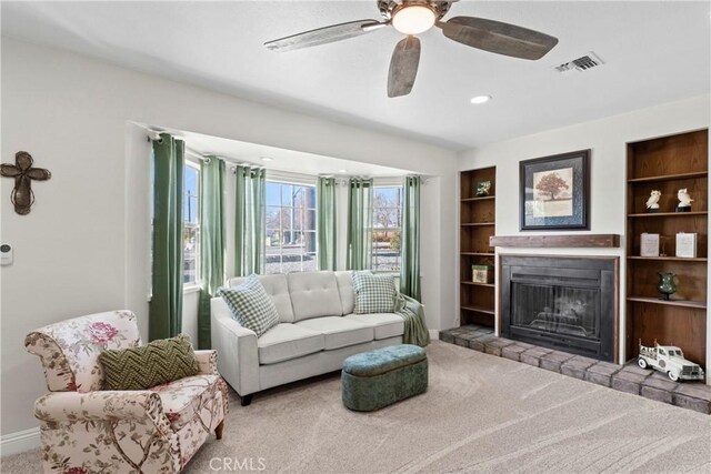 living area featuring built in shelves, visible vents, a glass covered fireplace, carpet flooring, and ceiling fan