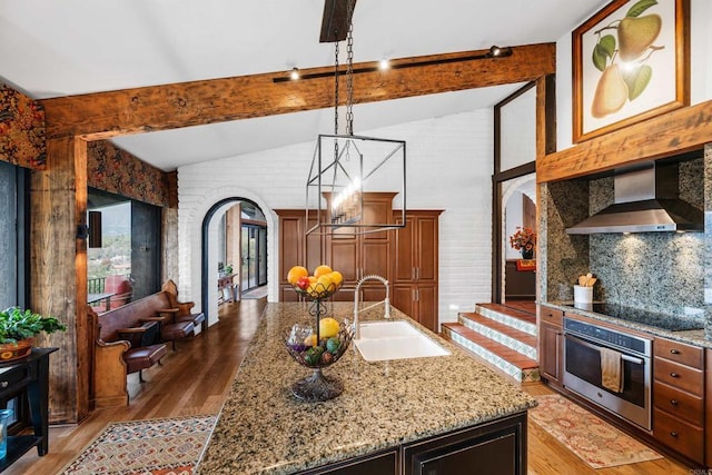 kitchen featuring sink, stainless steel oven, an island with sink, black electric stovetop, and wall chimney range hood