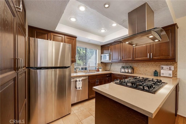 kitchen featuring stainless steel appliances, a sink, light countertops, a tray ceiling, and island exhaust hood