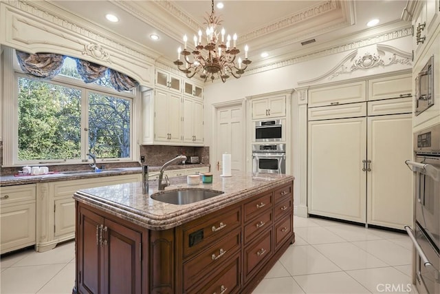 kitchen featuring sink, a raised ceiling, a kitchen island with sink, cream cabinets, and backsplash