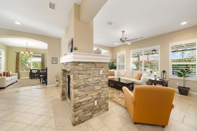 living room featuring light tile patterned flooring, a stone fireplace, and ceiling fan with notable chandelier