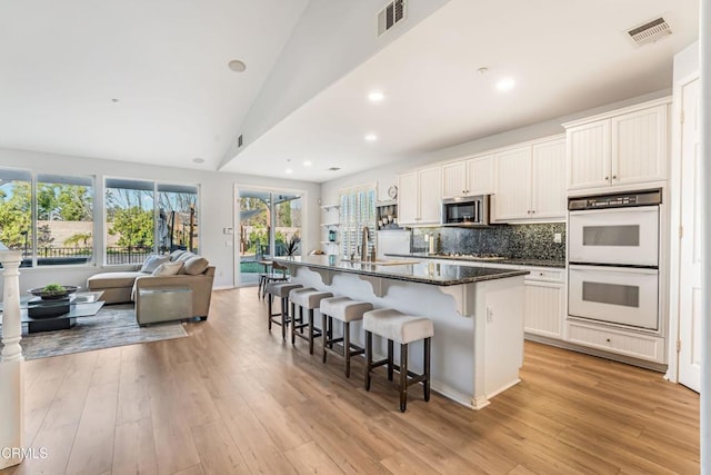 kitchen with double oven, white cabinetry, an island with sink, a breakfast bar area, and dark stone countertops