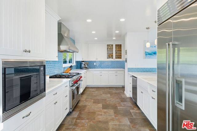 kitchen featuring white cabinets, hanging light fixtures, built in appliances, light stone countertops, and wall chimney range hood