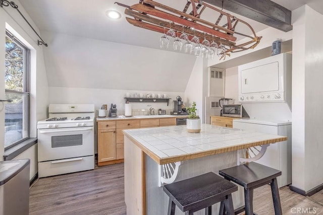 kitchen with a breakfast bar area, lofted ceiling with beams, tile counters, stacked washer / dryer, and white gas range oven