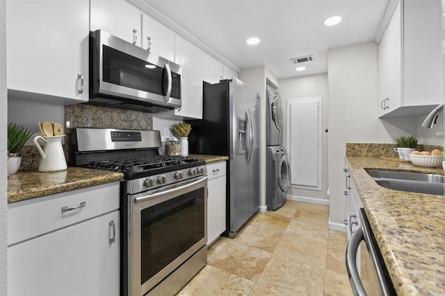 kitchen with sink, white cabinetry, stainless steel appliances, stacked washer / drying machine, and dark stone counters