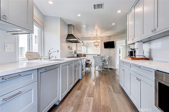 kitchen with pendant lighting, wall chimney range hood, sink, light hardwood / wood-style flooring, and appliances with stainless steel finishes