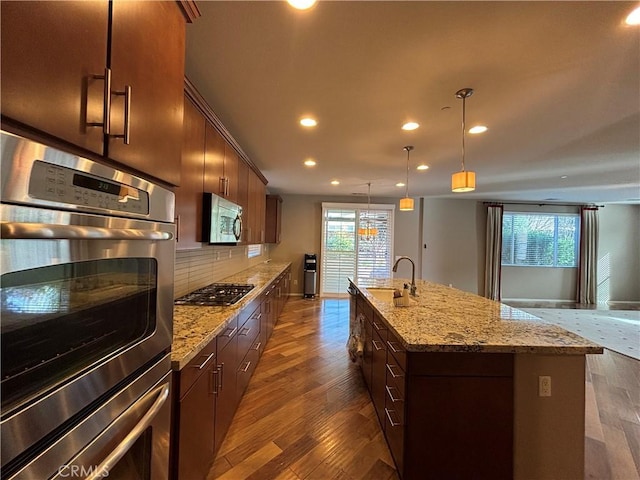 kitchen with appliances with stainless steel finishes, a kitchen island with sink, hanging light fixtures, dark hardwood / wood-style floors, and tasteful backsplash