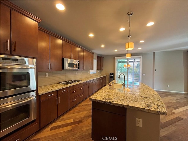 kitchen featuring dark wood-type flooring, appliances with stainless steel finishes, a kitchen island with sink, tasteful backsplash, and decorative light fixtures